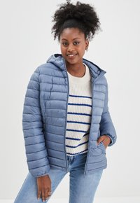 Lichtblauwe geïsoleerde jas met horizontaal doorgestikte patroon, aangehechte capuchon en ritssluiting, gedragen over een crèmekleurige gebreide trui met blauwe strepen.