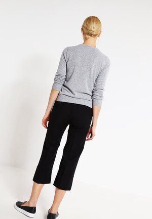 Kobieta o blond włosach związanych w niski kok, ubrana w szary top z długim rękawem, czarne spodnie do połowy łydki i czarne wsuwane buty, stojąca na tle białego tła.