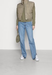 Dunjakke i lysebrun, grøn sweater, blå straight-leg jeans, hvide sneakers og en lille grøn håndtaske. Flad overflade som baggrund.