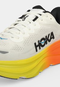 Λευκό και μαύρο αθλητικό παπούτσι Hoka με σόλα διαβάθμισης από κίτρινο σε πορτοκαλί και λευκά κορδόνια, παρουσιάζοντας πλαϊνές και μπροστινές λεπτομέρειες.