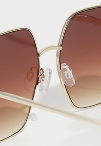 Ochelari de soare pătrați cu lentile gradiente de culoare brună și accente metalice aurii. Padelele nazale transparente, ajustabile, îmbunătățesc confortul și se potrivesc.