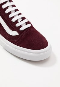 Gros plan d'une chaussure de sport en daim bordeaux avec des lacets blancs, une bande latérale blanche et une semelle en caoutchouc blanc épaisse et texturée sur fond blanc.