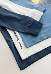 Fyra t-shirts staplade, med varierande nyanser av blått, vitt och marinblått. Mjukt bomullsmaterial med ribbade manschetter och tryckta etiketter.