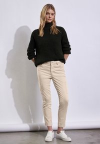 Un pull à col roulé tricoté épais de couleur vert foncé associé à un pantalon cargo beige avec poches latérales, porté avec des baskets blanches sur un fond gris.