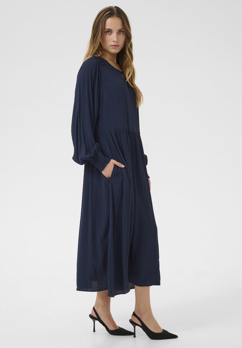 Maxikleid aus strukturiertem, marineblauem Stoff, mit langen Ärmeln, lockerem Schnitt und Seitentaschen. Kombiniert mit schwarzen High-Heels.