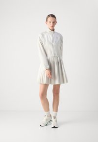 Vit klänning med plisserad kjol, långa ärmar, halvdragkedja vid kragen och dragsko i midjan. Bärs med vita sneakers och ankelsockor.