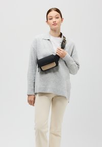 Czarna i beżowa torebka crossbody z zamkiem błyskawicznym, gładką teksturą i regulowanym paskiem, noszona na szarym swetrze i kremowych spodniach.