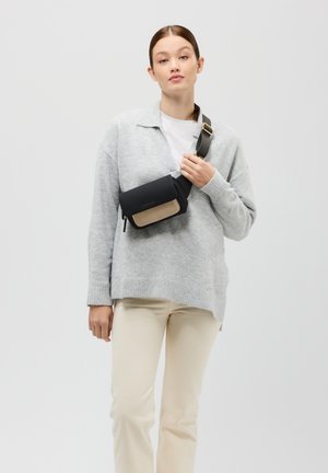 Borsa a tracolla nera e beige con chiusura a zip, texture liscia e tracolla regolabile, indossata sopra un maglione grigio e pantaloni crema.