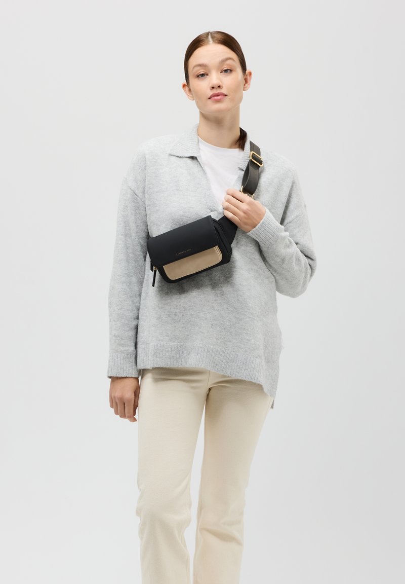 Czarna i beżowa torebka crossbody z zamkiem błyskawicznym, gładką teksturą i regulowanym paskiem, noszona na szarym swetrze i kremowych spodniach.