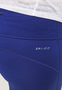 Närbild av blåa träningsleggar med "DRI-FIT" tryckt nära midjan, i kombination med en vit topp som visar en svag "DRI-FIT" logotyp.