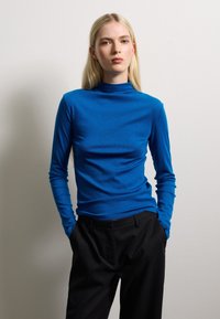 Blue turtleneck top, long sleeves, slim fit, smooth texture, paired with black tailored pants, against a neutral background.