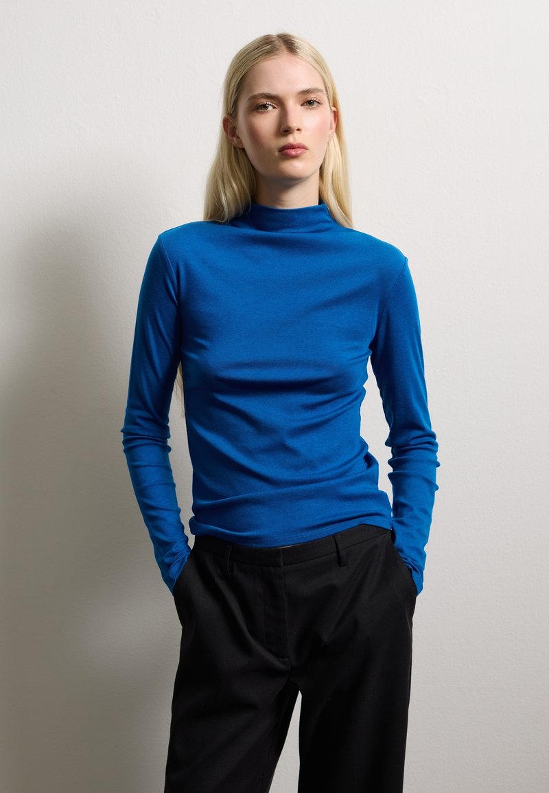 Blue turtleneck top, long sleeves, slim fit, smooth texture, paired with black tailored pants, against a neutral background.
