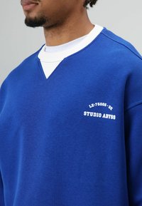 Blauwe trui met een V-hals en een witte binnenkraag, met de opdruk "LE-75005-DE" en "STUDIO ABYSS" in het wit. Zachte textuur.