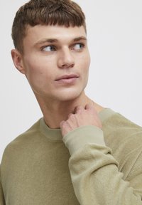 Olivenfarget sweatshirt med en teksturert overflate, ribbestrikket krage og mansjetter, med lange ermer og avslappet passform, som fremhever mykt materiale.