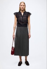 Fekete áttetsző blúz fodros ujjakkal és gombos részlettel, hosszú szürke szoknyával párosítva. Egy kis bordó táskát tart a kezében. Fekete cipő.