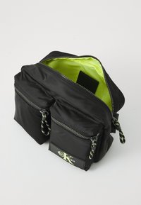 Black nylon bag with dual zippered compartments, neon green interior lining, and textured pull tabs featuring a visible logo patch.