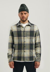 Geruiten shirt in groen, beige en marineblauw, met een knoopsluiting aan de voorkant, een borstzak en lange mouwen. Gecombineerd met een groene beanie.