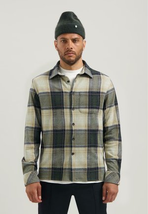 Geruiten shirt in groen, beige en marineblauw, met een knoopsluiting aan de voorkant, een borstzak en lange mouwen. Gecombineerd met een groene beanie.