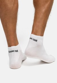 Hvide ankelsokker lavet af en tekstureret bomuldsblanding, med et ensfarvet design og "INDICODE"-logo i sort på hælen.