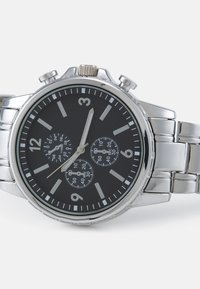 Montre en acier inoxydable argenté avec un cadran noir, des fonctionnalités de chronographe et des index horaires luminescents. Elle possède un bracelet à maillons métalliques et une couronne texturée.