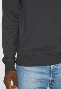 Hombre vistiendo un sudadera de manga larga gris oscuro y jeans de mezclilla azul claro, con la mano relajada a un lado contra un fondo liso.