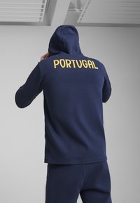 Jūrinis kapučino su gobtuvu ir geltonu tekstu, skaitančiu "PORTUGALIJA" nugaros pusėje. Pagamintas iš minkštos audinio, turi laisvą kirpimą ir minimalų dekorą.