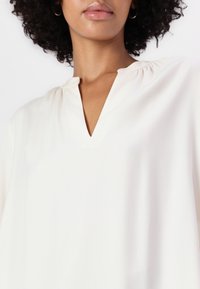 Lys ivorfarget bluse med en myk tekstur, har V-hals og rynkedetaljer ved kragen, og viser en avslappet passform og minimalistisk design.