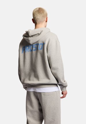 Osoba z krótkimi blond włosami, ubrana w jasnoszary kaptur i dopasowane spodnie dresowe, na plecach kaptura duży niebieski napis "PROSTO".