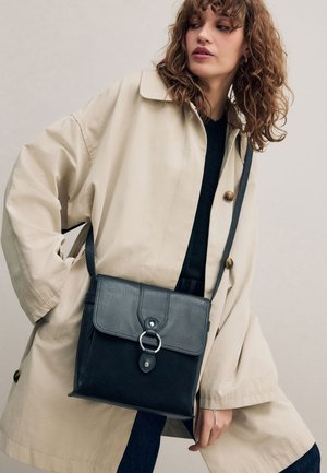 Femeie purtând un trench bej și un top negru, ținând o geantă crossbody din piele neagră cu detaliu de inel argintiu.