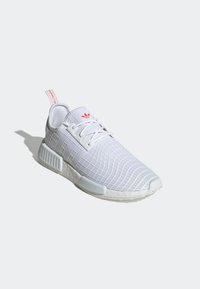 Zapatilla deportiva blanca con patrón de cuadrícula texturizado, parte superior de malla ligera, suela de goma y acento rojo en la lengüeta y la pestaña del talón.