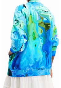 Osoba nosící výraznou modrozelenou mikinu s abstraktním vzorem, rukávy vyhrnuté, a bílou sukni, viděná zezadu.