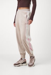 Osoba oblečená v béžových sametových teplácích Adidas s růžovým logem na straně, v hnědém zkráceném svetru s dlouhým rukávem a béžových teniskách Adidas.