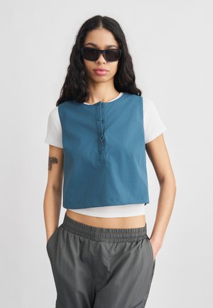 Kvinde med langt mørkt hår iført sorte solbriller, blå ærmeløs top over hvid kort skjorte og grå bukser med elastik i taljen, stående med hænderne i lommerne.