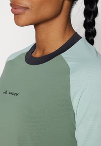 Grønn og lyseblå kortermet skjorte med svart krage, laget av et teksturert, pustende stoff, med Vaude-logoen på fronten.