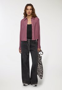 Jeans scuri e larghi abbinati a una camicia cropped button-up color malva. La modella tiene in mano una borsa a tracolla a righe zebra in bianco e nero.