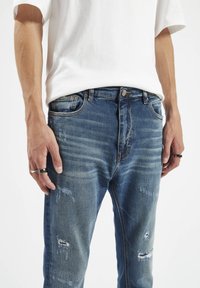 Vaqueros de denim azul deslavado con detalles desgastados, que presentan un ajuste slim, cinco bolsillos y costuras en contraste. Llevados con una camiseta blanca.