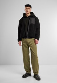 Chaqueta de forro polar negra con un panel negro en contraste, llevada sobre una sudadera negra; combinada con pantalones caqui y botas negras gruesas.