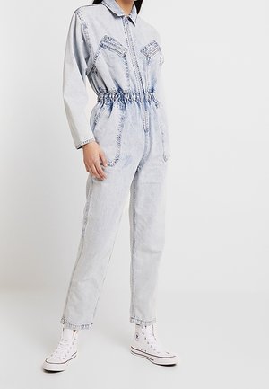 Nainen, joka on pukeutunut vaaleansiniseen happopesulla käsiteltyyn farkkuhaalariin, jossa on joustava vyötärö, vetoketju edessä sekä rinta- ja reisitaskut, yhdistettynä valkoisiin korkeavartisiin tennareihin.