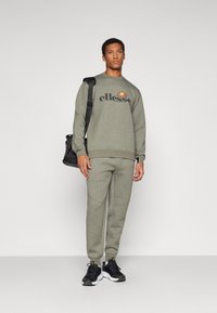 Zelena majica s kapuljačom s logotipom "ellesse", u kombinaciji s odgovarajućim uskim trenirkama. Ima rebraste manžete, bočne džepove i crne sportske cipele.