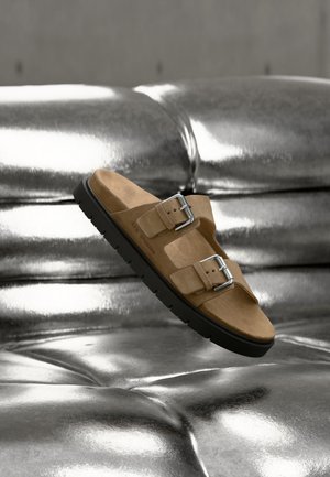 Braune Wildledersandale mit zwei silbernen Schnallen, die über einer glänzenden silbernen, gesteppte Oberfläche schwebt, mit schwarzen Sohlen- und Fußbett-Details.