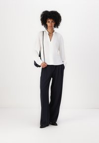 Blusa bianca a maniche lunghe con scollo a V e dettagli con bottoni, abbinata a pantaloni larghi blu navy e una borsa a tracolla nera. Scarpe nere.