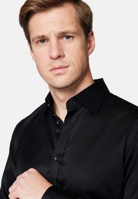 Czarna koszula zapinana na guziki z gładkiego materiału z eleganckim kołnierzykiem, o dopasowanym kroju i subtelnym połysku, pokazana z ręką modela na kołnierzyku.