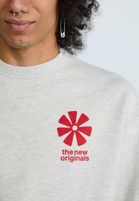 Šviesiai pilkas džemperis su raukšlėta apykakle, su raudona gėlių logotipu ir tekstu "nauji originalai" žemiau. Minkšta, tekstūruota medžiaga.
