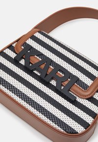 Vävd handväska i svart och vitt med ränder, med bruna läderdetaljer och en framträdande svart logotyp på framsidan. Fyrkantig form och justerbar rem.