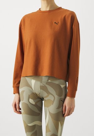 Woman wearing ribbed burnt orange long-sleeve shirt with Puma logo and camo-patterned leggings against white background.