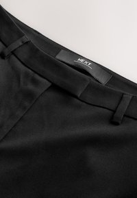 Svart tygmidjeband med en V-formad skärning, med en sydd etikett där det står "NEXT", som visar en slät textur och skräddarsydda stängningsdetaljer.