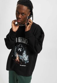 Melna liel izmēra sporta krekls ar grafiku, kurā attēlots lācis un koki, ar uzrakstu "bet aukstā sirds". Modelis ģērbies ķēdes kaklarotā un zaļās biksēs.