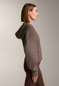 Bruine hoodie met een losse pasvorm, zachte textuur en een capuchon. Geribbelde boorden aan de mouwen en zoom; gecombineerd met donkerbruine leggings. Zijaanzicht.