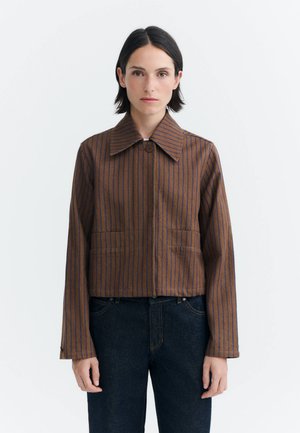 Femme aux cheveux courts et foncés portant une veste marron avec de fines rayures verticales bleues et un jean en denim foncé, debout devant un fond clair uni.