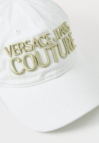 Bela kapa z višinim zlatim vezenjem z napisom "VERSACE JEANS COUTURE." Narejena je iz trpežnega bombažnega blaga, s ukrivljenim vizirjem in strukturiranim dizajnom.